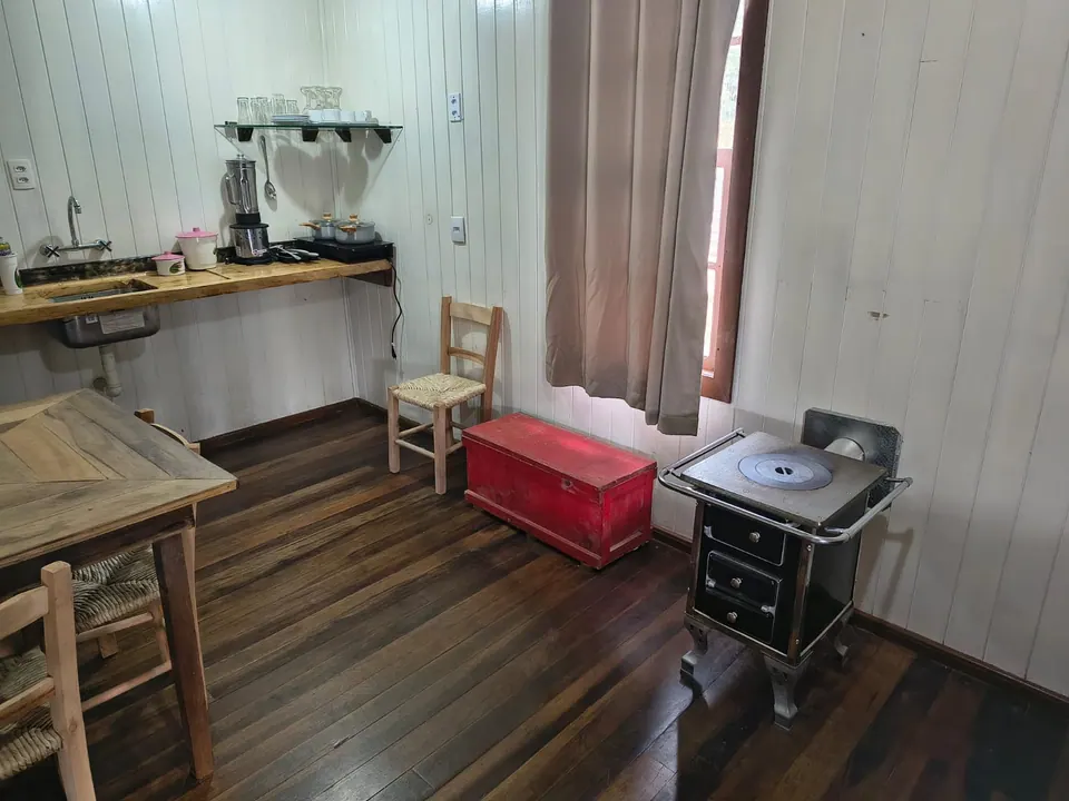 Cozinha com fogão a lenha, bancada de madeira, mesa rústica, cadeira de palha e baú vermelho